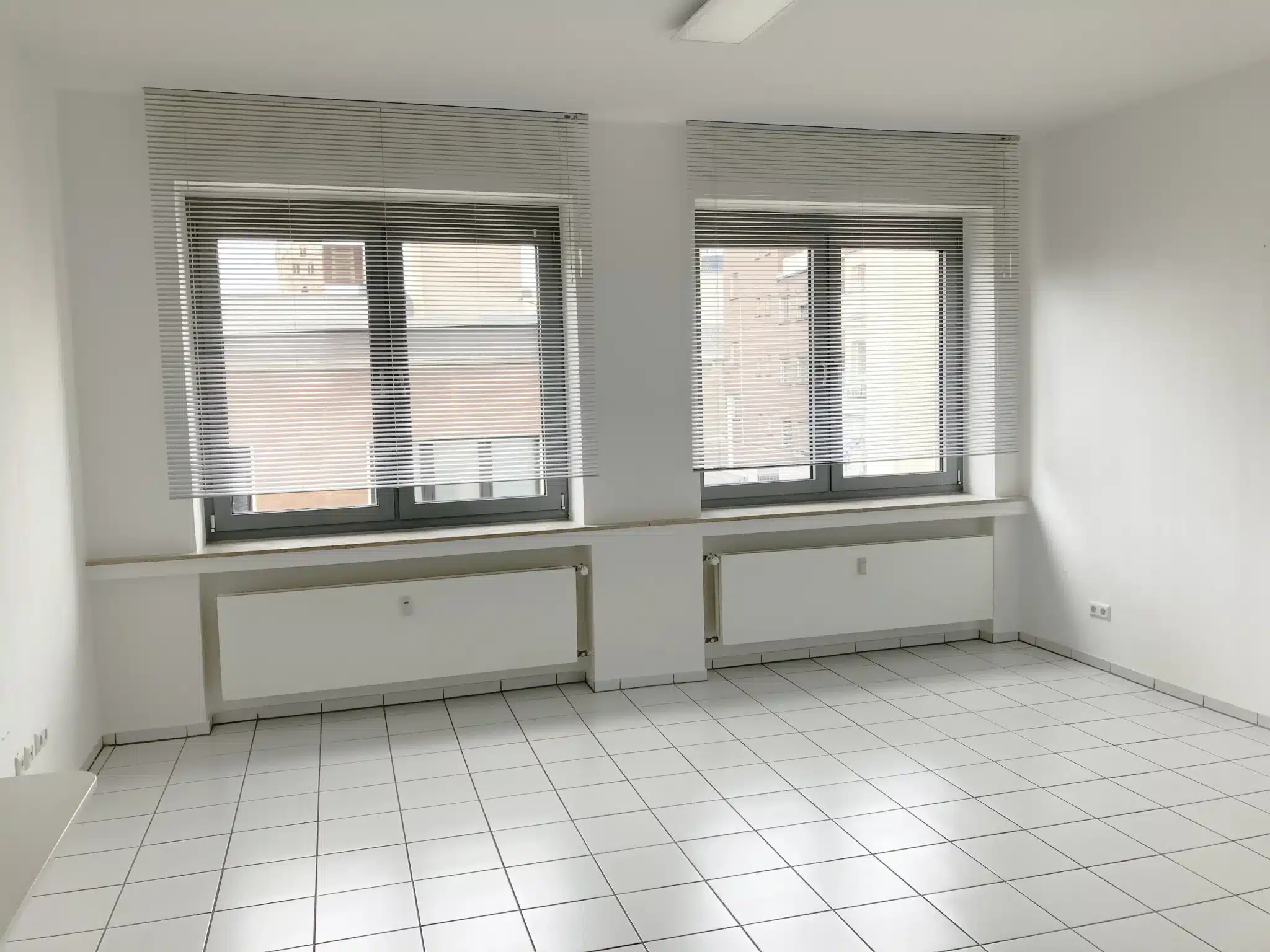 Heller leerer Büroraum mit großen Fenstern und weiß gefliestem Boden.